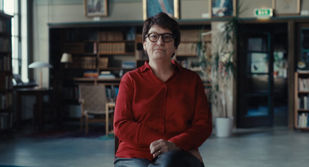 Helle Klein sitter på en pall i ett bibliotek och tittar in i kameran.