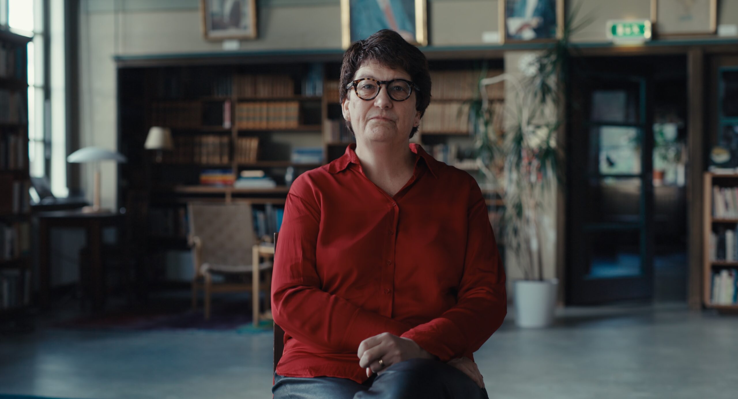 Helle Klein sitter på en pall i ett bibliotek och tittar in i kameran.