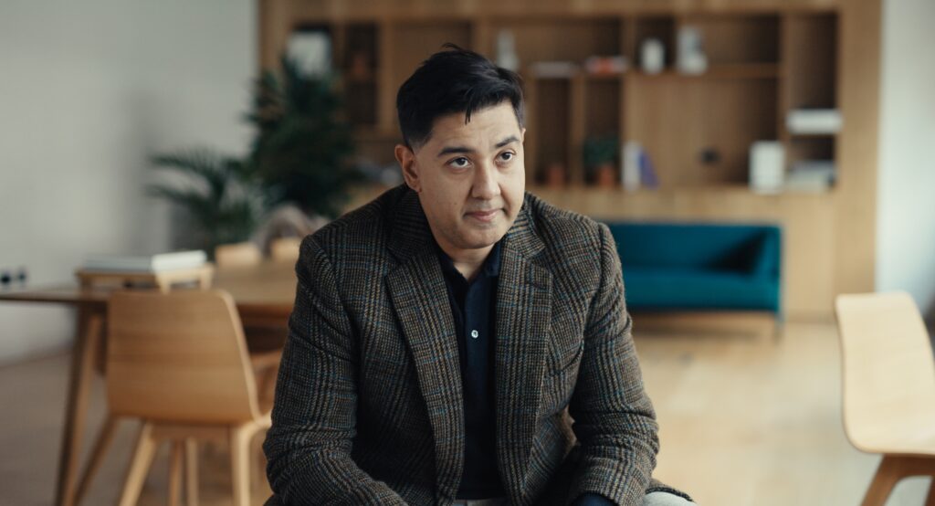 Imran Ahmed sitter på en pall i ett tomt mötesrum. Han har mörkthår, kavaj och svart skjorta.