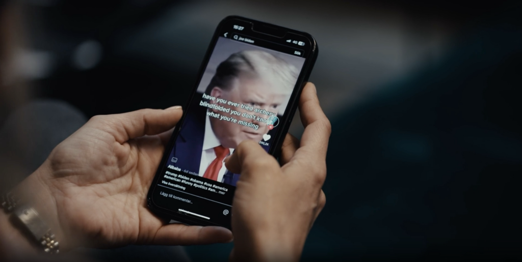 Två händer håller en mobiltelefon, på skärmen syns en bild av Donald Trump.